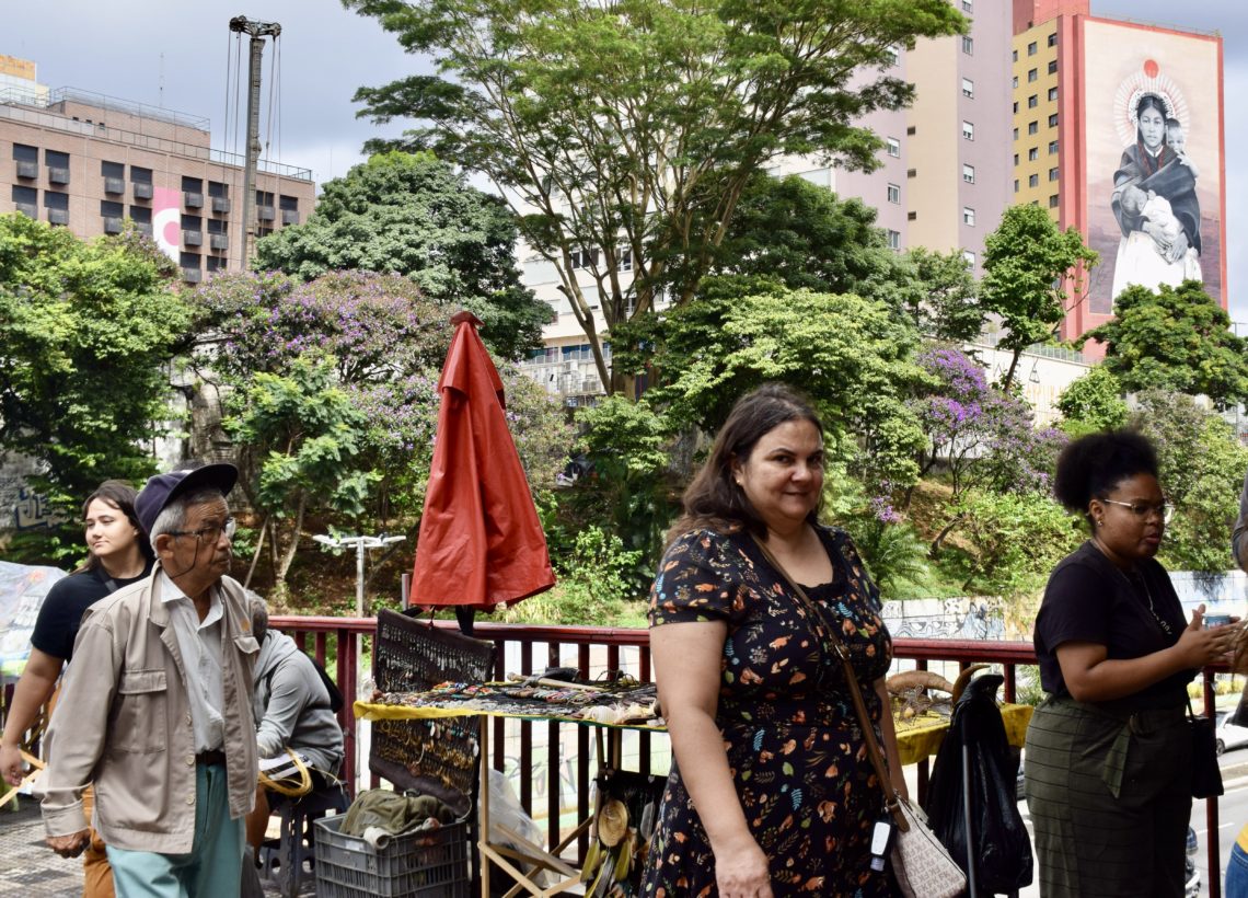 Personas caminando y conversando en un mercado callejero en São Paulo, con un gran mural al fondo