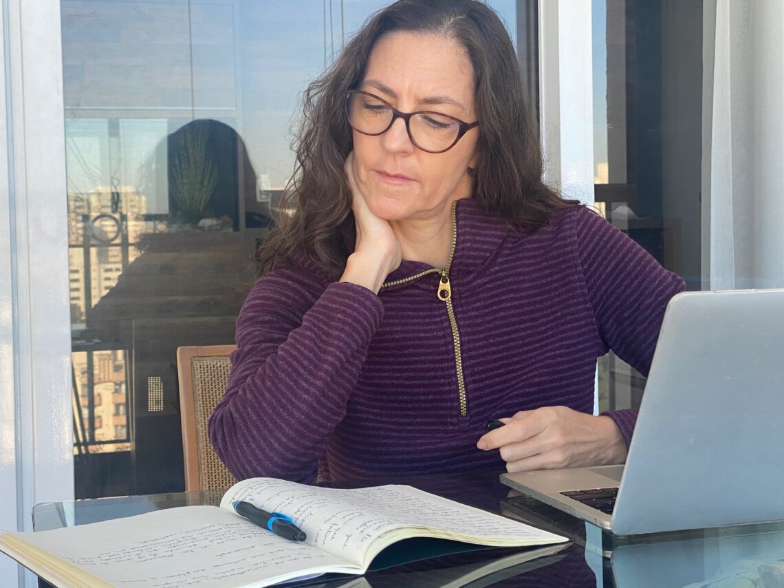 Claudia Mendizábal escribiendo en un cuaderno frente a su computadora portátil, sentada en un balcón con vista urbana