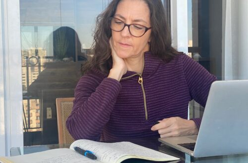 Claudia Mendizábal escribiendo en un cuaderno frente a su computadora portátil, sentada en un balcón con vista urbana