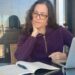 Claudia Mendizábal escribiendo en un cuaderno frente a su computadora portátil, sentada en un balcón con vista urbana