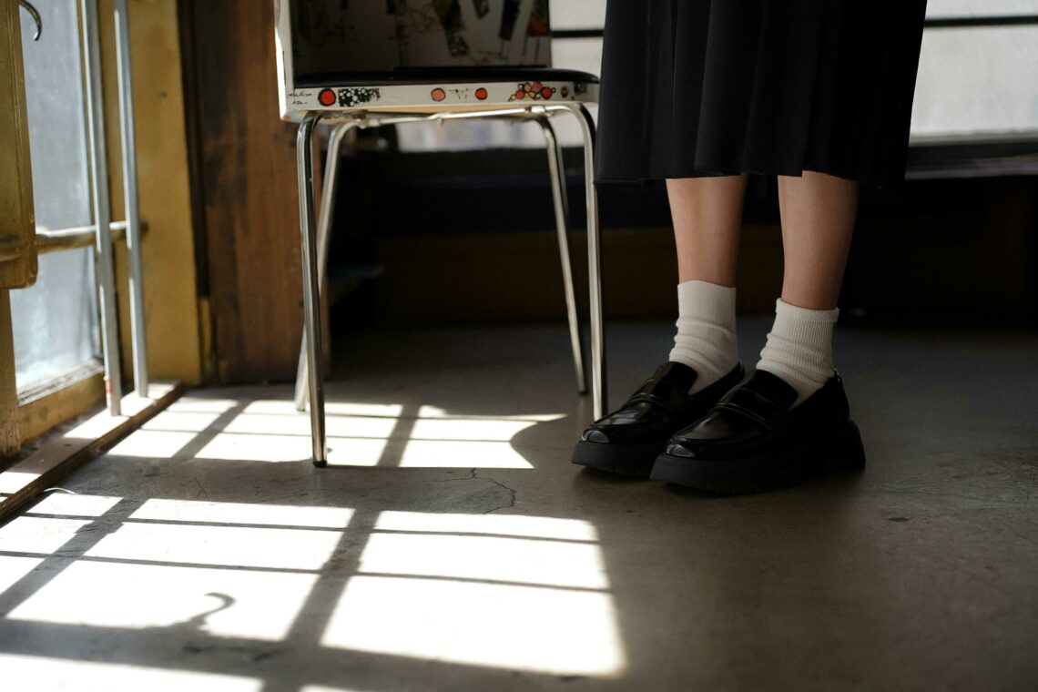 Piernas con medias y zapatos negros detrás de una silla metálica, luz entrando por la ventana