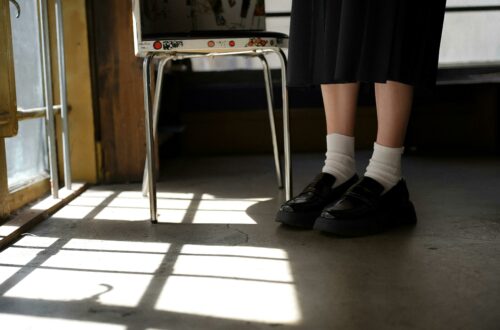Piernas con medias y zapatos negros detrás de una silla metálica, luz entrando por la ventana