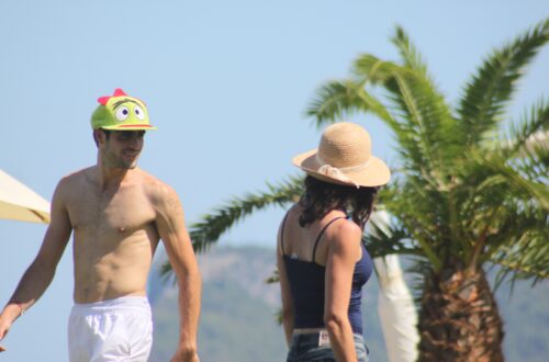 Hombre sonriente con gorra verde divertida y mujer con sombrero de paja caminando bajo palmeras en un día soleado.