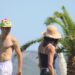 Hombre sonriente con gorra verde divertida y mujer con sombrero de paja caminando bajo palmeras en un día soleado.